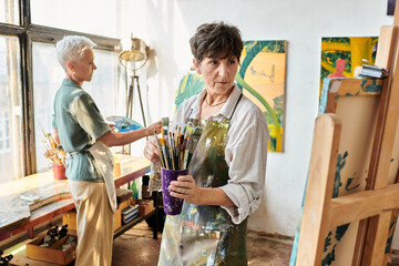talented mature woman with paintbrushes looking at easel near female friend in art workshop