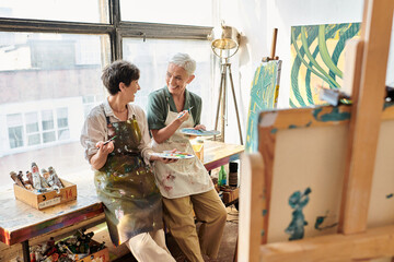 excited mature female artists with palettes and paintbrushes talking near easel in craft workshop