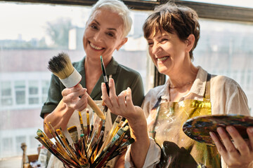 cheerful mature female women holding different paintbrushes during master class in art studio