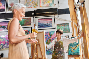 Fototapeta na wymiar smiling mature model with wine glasses posing near woman artist in art studio, creative process