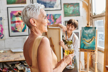 joyful and stylish woman posing with wine glass near female artist in workshop, creative process