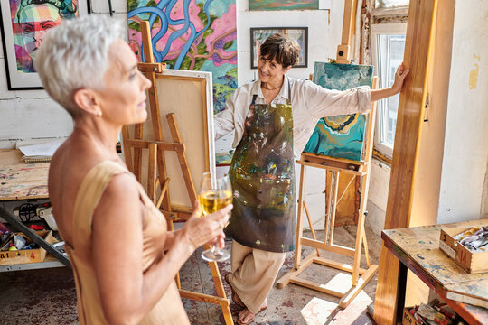happy woman artist looking at elegant middle aged model posing with wine glass in art workshop