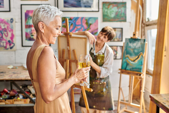 happy and elegant woman posing with wine glass near female artist in workshop, creative process