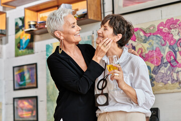 trendy mature woman touching face of lesbian partner with wine glass in contemporary art studio