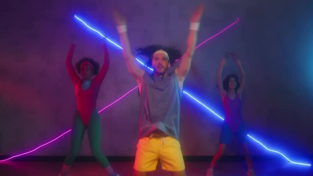 Medium long shot of group of biracial sportspeople doing jumping jacks while practicing aerobics in retro activewear in studio with purple neon light