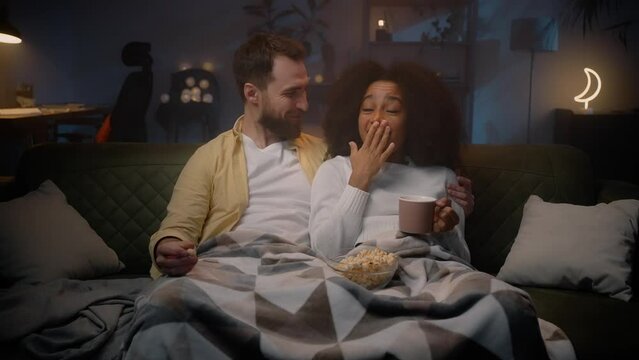 Multiracial spouses sitting on couch and spending weekend with popcorn and movie