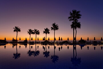 Morgendämmerung am Pool mit Spiegelung auf dem Wasser, Palmen und Sonnenschirme im Hintergrund
