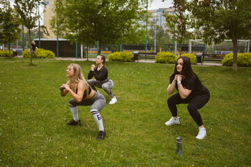 Fototapeta premium Happy female athletes do squat exercises in morning square. Young women do cardio training to refine figure. Healthy habits concept