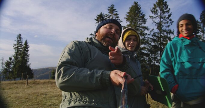 Caucasian man records video with friends or family on the top of the hill and talks on smartphone front camera. Group of diverse travelers during hike in the mountains. POV video call or filming vlog.