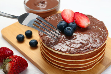 Eating tasty pancakes with chocolate paste and berries with fork on white wooden table, closeup
