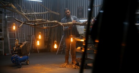 African American carpenter ties with rope design wood product from tree branch with light bulbs. Artisan or designer woodworking in modern garage or workshop. Hand craft and entrepreneurship concept.