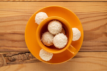 Several candies with nut and coconut flakes with ceramic saucer and cup on wooden table, macro, top view.