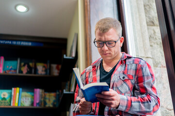 male professor in glasses sits in the library and carefully reads a book on the background of...