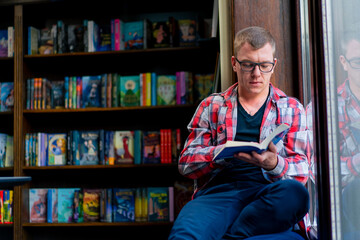 male professor in glasses sits in the library and carefully reads a book on the background of...