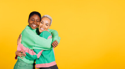 Two young cheerful women wearing green clothes embracing in a studio with a yellow background.