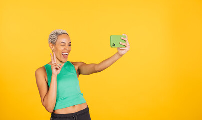 Happy young woman taking a selfie in front of a yellow background in a studio. Woman taking a...