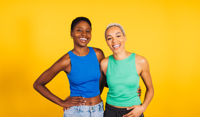 Portrait of a young cheerful female couple in front of a yellow background at studio. Two women...