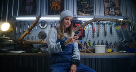 Female carpenter, craftswoman uses mobile phone sitting in stylish workshop. Wood product made from tree branch and board with professional tools in the background. Handicraft and entrepreneurship.