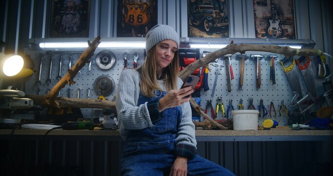 Female carpenter, craftswoman uses mobile phone sitting in stylish workshop. Wood product made from tree branch and board with professional tools in the background. Handicraft and entrepreneurship. - Powered by Adobe