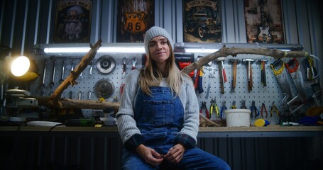 Female carpenter, artisan smiles and looks at camera sitting in modern stylish workshop. Wooden product from tree branch and board with professional tools in background. Handmade and small business.