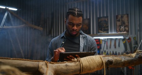 Creative African American designer uses digital tablet computer while creating design wooden products in modern garage. Craftsman or carpenter works in stylish workshop. Handmade and entrepreneurship.