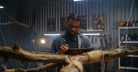 Creative African American designer uses digital tablet computer while creating design wooden products in modern garage. Craftsman or carpenter works in stylish workshop. Handmade and entrepreneurship.