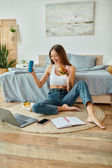 happy appealing woman in casual wear sitting on floor and eating burger while working on her laptop