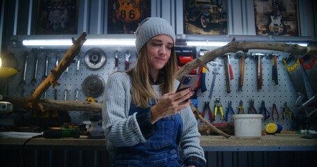 Female carpenter, craftswoman uses mobile phone sitting in stylish workshop. Wood product made from tree branch and board with professional tools in the background. Handicraft and entrepreneurship.