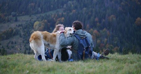Caucasian couple of hikers sits on the hill, takes photos on phone, pets and plays with local dog....