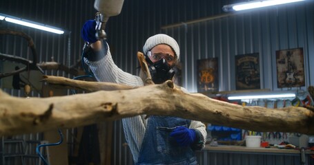 Woman artisan wearing respirator sprays liquid or wood varnish on tree branch. Creative female carpenter woodworking, creates wooden products in stylish workshop. Handmade and small business concept.