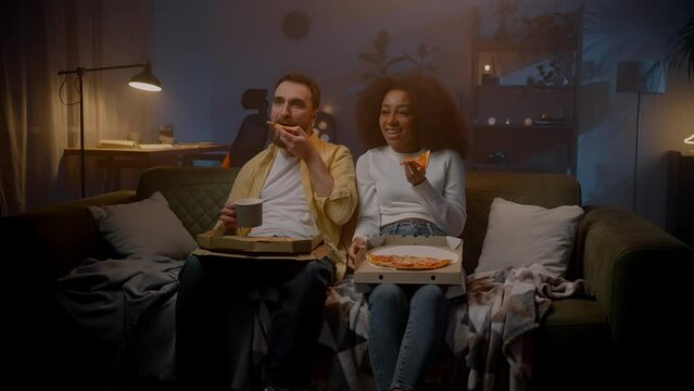 Multiracial spouses sitting on couch and spending weekend with pizza and comedy movie