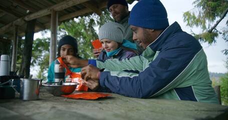 Adult African American man opens doy pack for his wife. Multiethnic tourist family eat hiking food together. Group of travelers or hikers rest in gazebo after expedition or trek in the mountains.