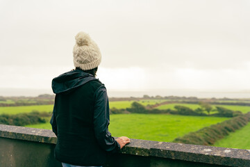 Unknown girl with wool hat standing by a railing to enjoy the Irish landscape on her road trip...