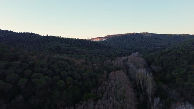 Vista a&eacute;rea de monta&ntilde;as al atardecer con bosques y una cantera iluminada a lo lejos. Bosque oscuro atardeciendo.