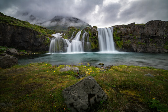 Lush green landscape with a distinctive mountain peak towering over dual waterfalls under a dramatic sky in Iceland - Powered by Adobe