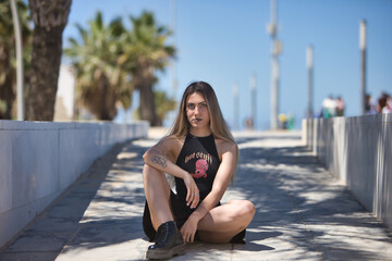 Young, beautiful blonde woman, green eyes, with black top and skirt, tattoos, sitting on the floor, looking at the camera, rebellious and independent. Concept looks, rebelliousness, insubordination.