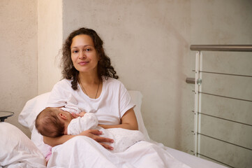 Beautiful Latin American woman, young loving caring mother in white pajamas, breastfeeding her baby...