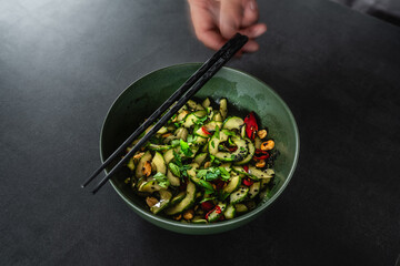 Asiatischer Gurkensalat auf dunklem Hintergund in dunkler Schale mit Essstäbchen und Hand