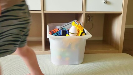 The child puts the toys in the box after playing, cleaning the room. Baby aged two years (two year old boy)