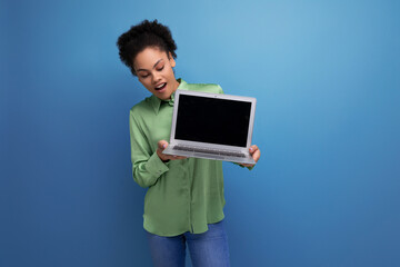 young pretty female model with curly black hair dressed in a green blouse and jeans holds a laptop screen forward with a mock up for advertising
