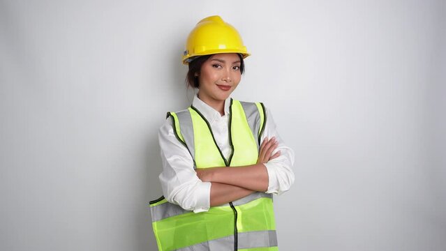 Smiling Asian woman labor worker in industry factory, posing with arms folded, wearing yellow safety helmet, green vest and uniform, isolated white background.