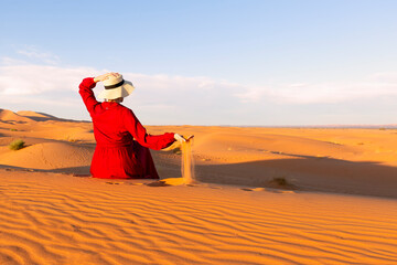 A young woman dressed in red and hat sitting on the desert dunes, playing with the sand - obrazy, fototapety, plakaty