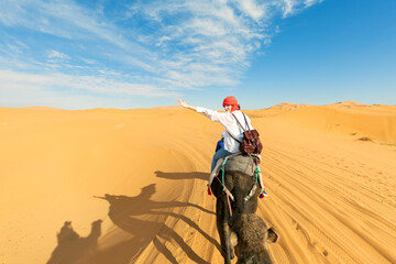 A young women  rides a camel through the dunes in the Sahara Desert. View of the woman from behind,...