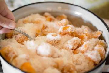 A woman stirs Apricots covered with sugar in an enamel pan to prepare fruit jam