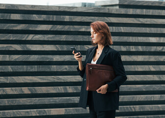 Side view of middle-aged businesswoman with leather folder and smartphone walking outdoors