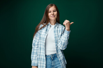 Charming young woman with brown hair points her hand to the side on a green background with copy space