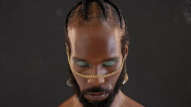 Black gay stands isolated on black background stares into camera with serious expression on face. Close-up portrait of handsome adult transgender with blue eyeshadows and golden accessory on face