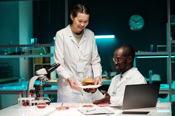 Asian scientist giving plate with hamburger with lab-grown meat to her black colleague