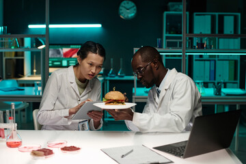 Two scientists analyzing hamburger with lab-grown meat, female researcher taking notes on tablet