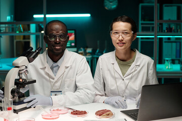 Two young biracial researchers posing together at desk in laboratory looking at camera